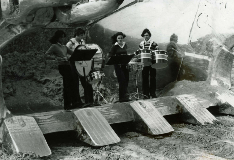 Four musicians playing instruments, performing in front of a concrete structure. Legacy