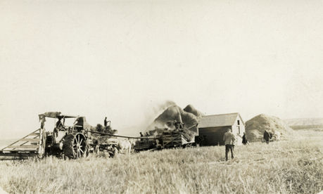 Vintage tractor and workers harvesting crops near a small building and stacks of hay.